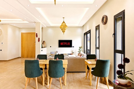 A modern hotel room with a wooden dining table, green upholstered chairs, a large window, and a wall-mounted TV displaying 'NETFLIX'.. Riad Villa Alain