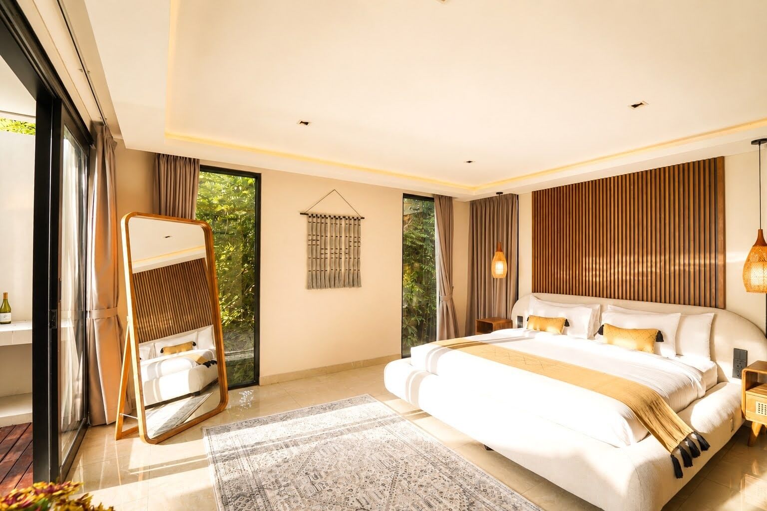A modern bedroom with a large bed, a mirror, a hanging light fixture, and a view of greenery outside.