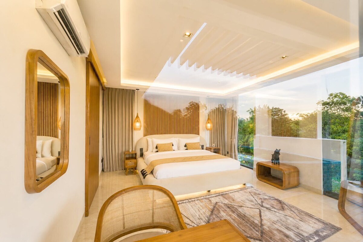 Ein modernes Schlafzimmer mit einem großen Bett, einem Holzstuhl, einem kleinen Tisch und Blick auf Grün durch große Fenster.