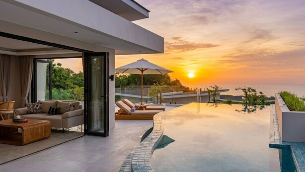 En modern infinitypool med utsikt över solnedgången, en soffa med kuddar och ett soffbord i trä.