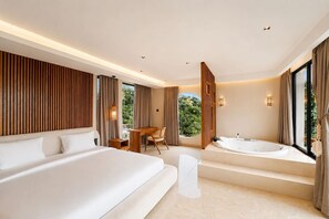 A modern hotel room with a large bed, a desk, a chair, a bathtub, and a view of greenery. - Zen Hillside Oceanview Bali (Badung)