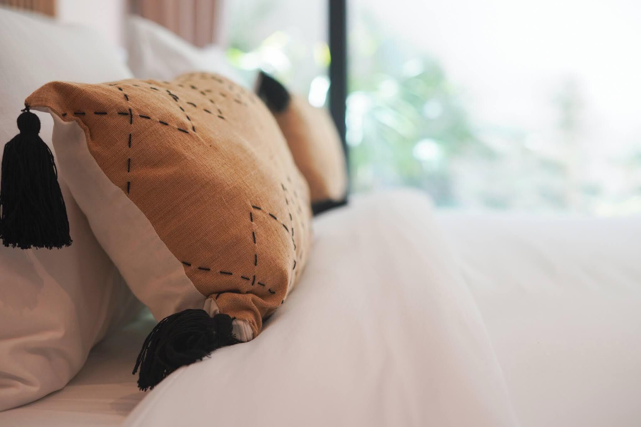 Una cama con sábana blanca y almohada color beige con flecos.