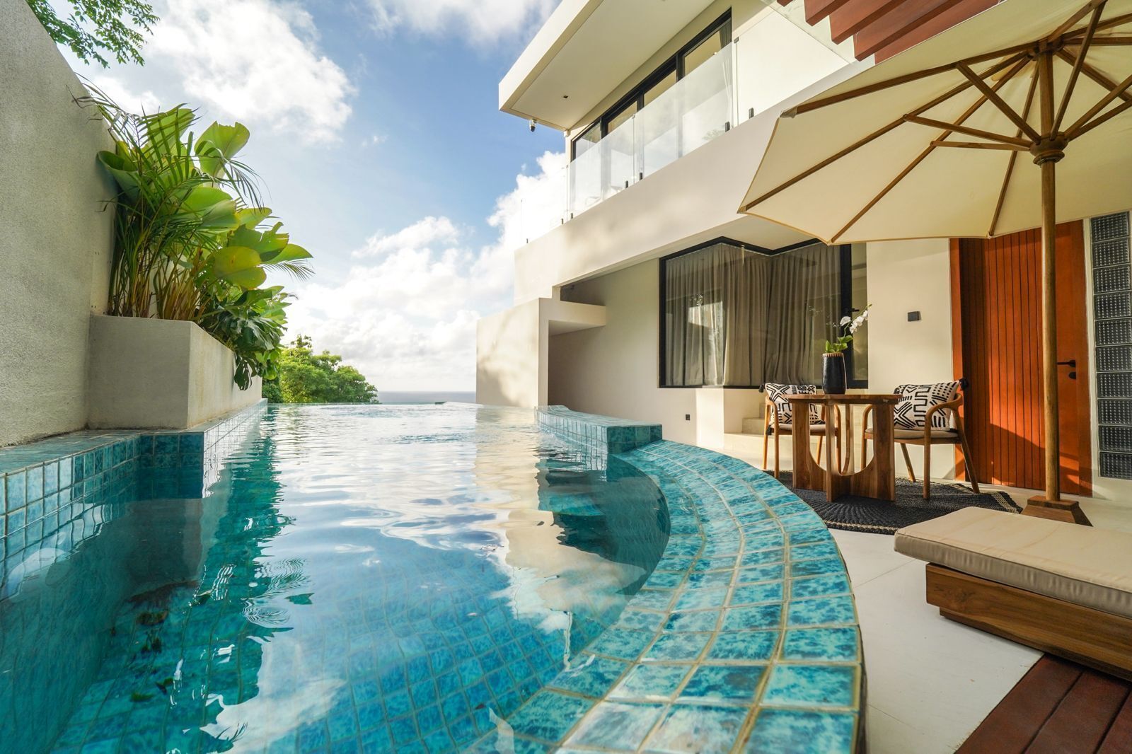 A infinity pool with a view of the ocean, a wooden deck, and a dining area with outdoor furniture.