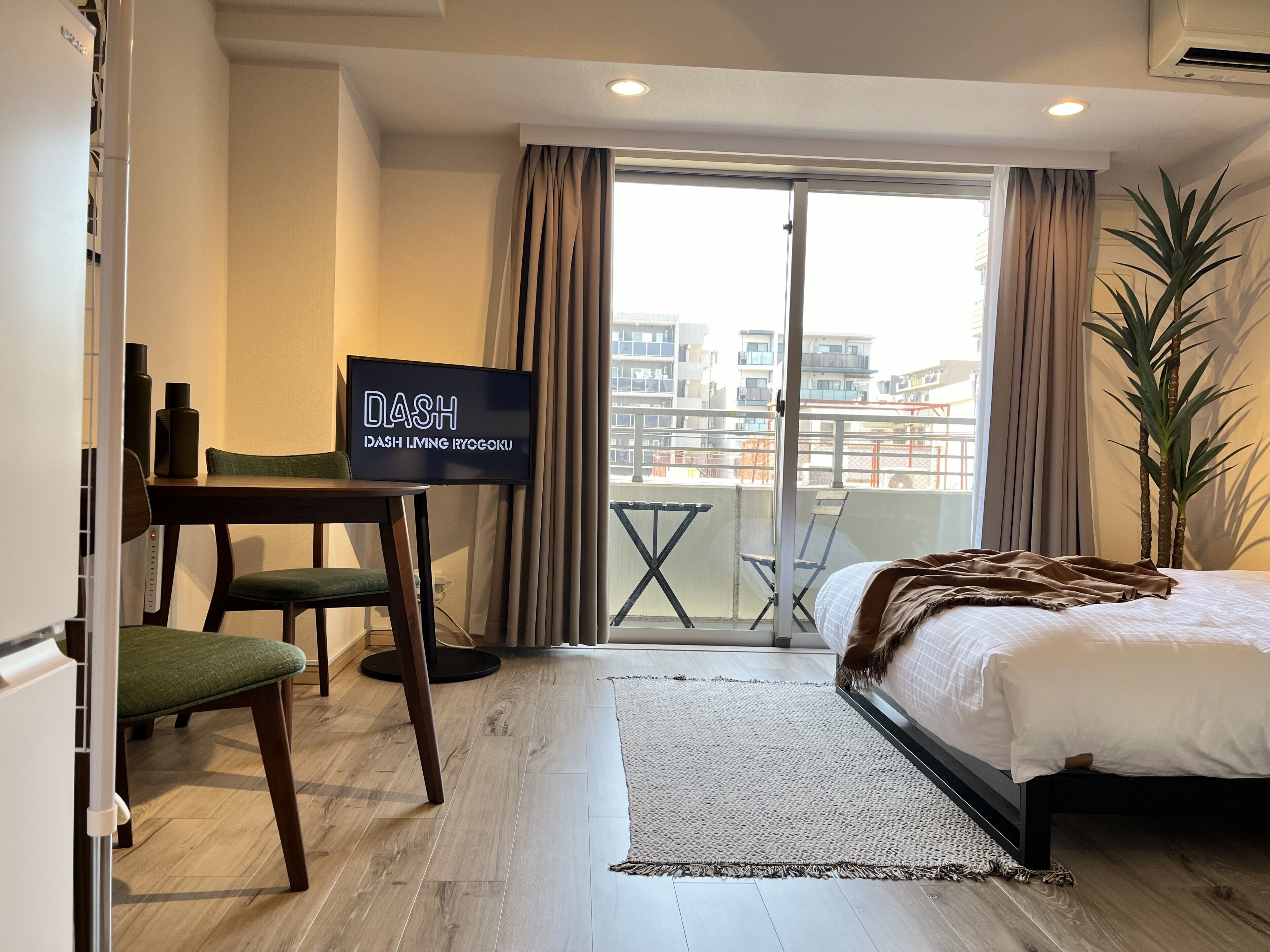 A hotel room with a bed, a TV, a desk with a chair, a balcony with a view of buildings, and a potted plant.