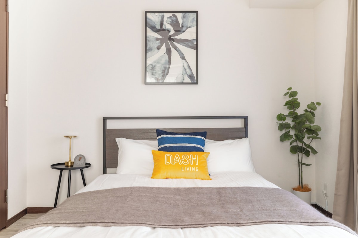 A neatly made bed with a gray blanket, a bedside table with a lamp, and a wall-mounted abstract painting.