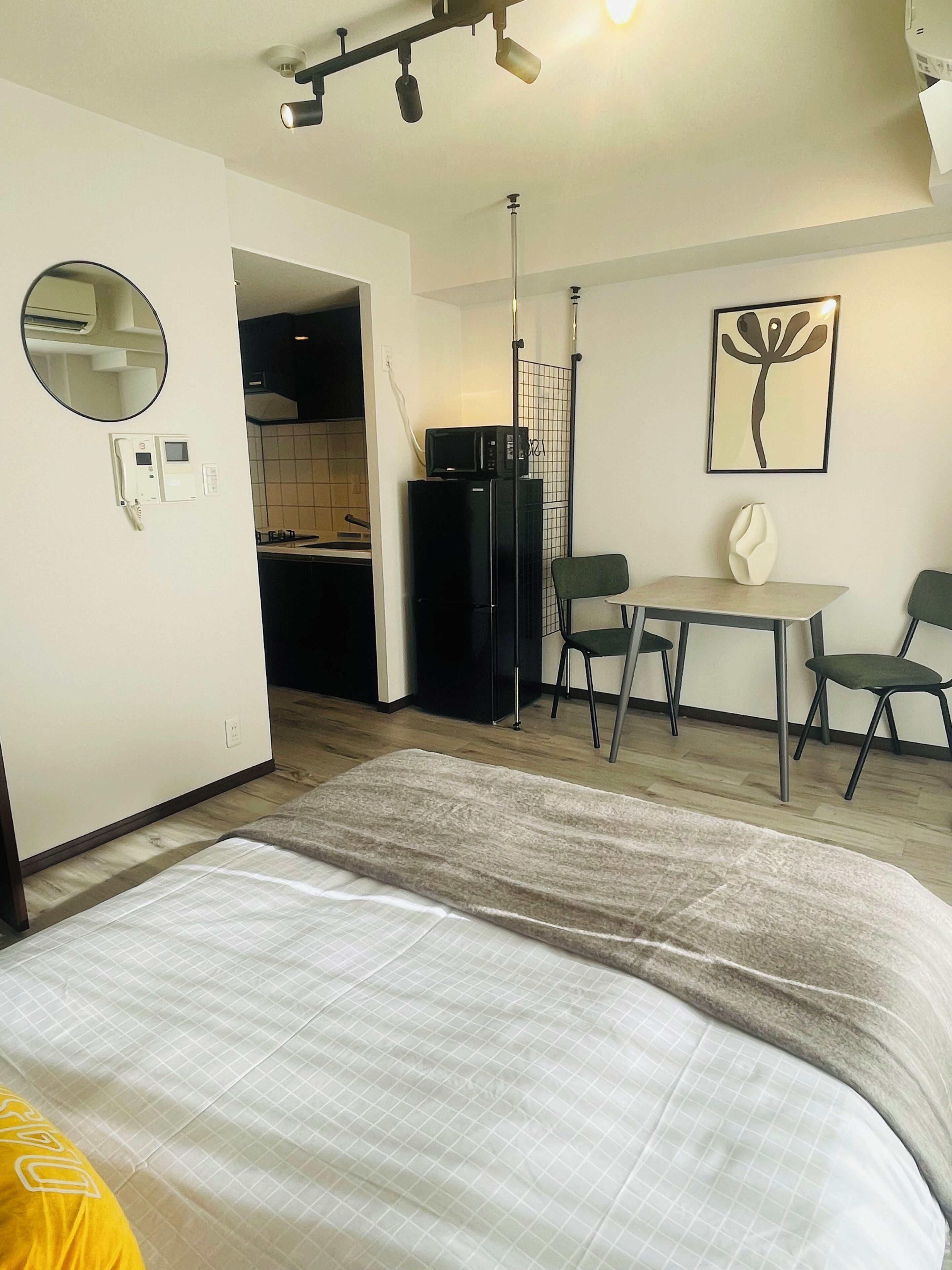 A compact hotel room with a bed, a small kitchenette, a dining table with chairs, a mirror, and a framed artwork on the wall.
