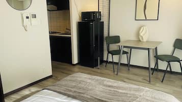 A compact hotel room with a bed, a small kitchenette, a dining table with chairs, a mirror, and a framed artwork on the wall.
