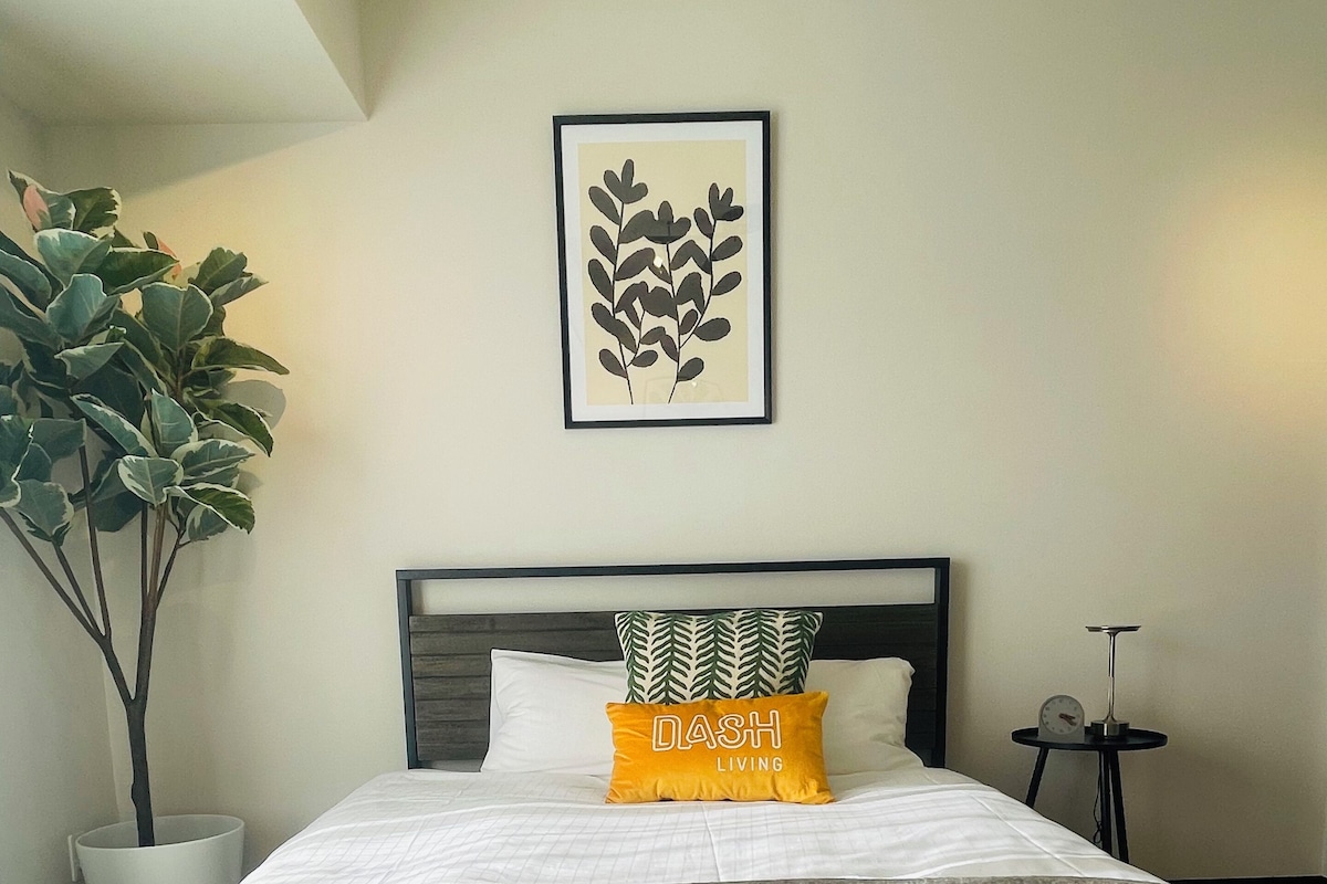 A neatly made bed with a gray blanket, a green and white patterned pillow, and a yellow pillow with the text 'DASH LIVING'.