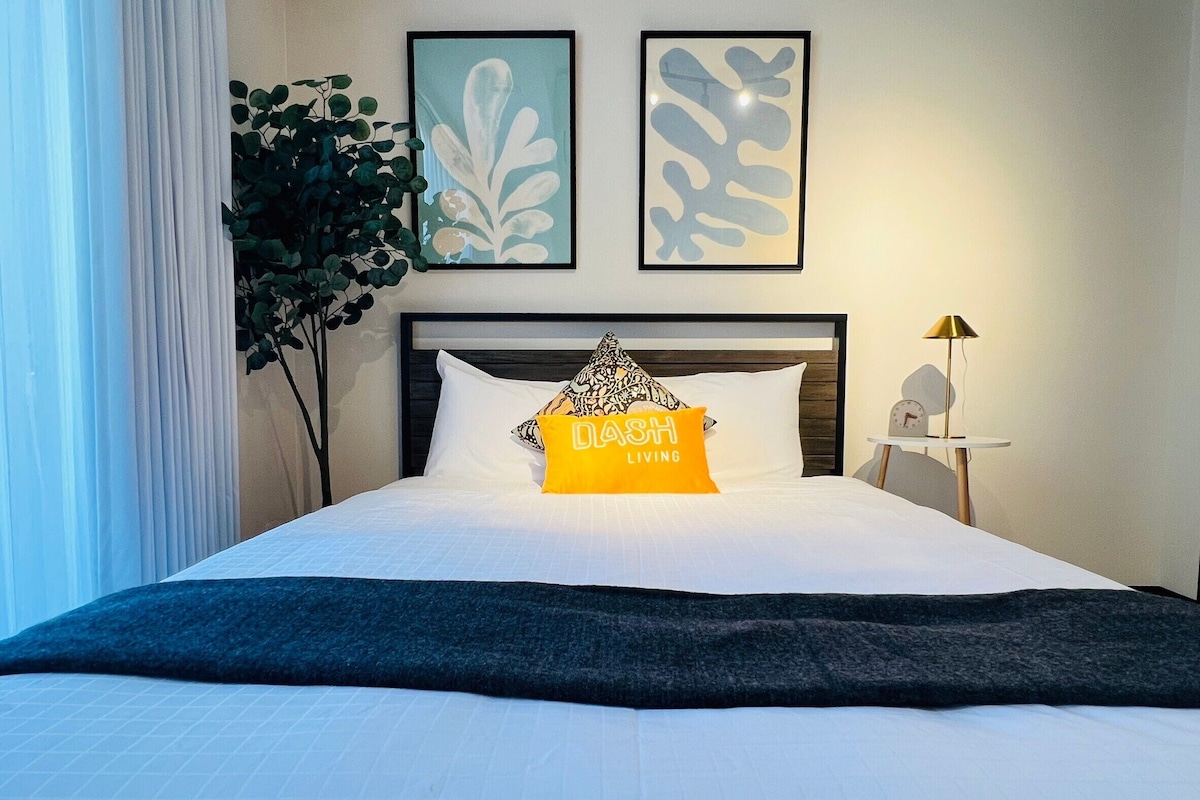A neatly made bed with white bedding, a black headboard, and a single yellow pillow with text. There are two framed abstract art pieces on the wall above the bed.