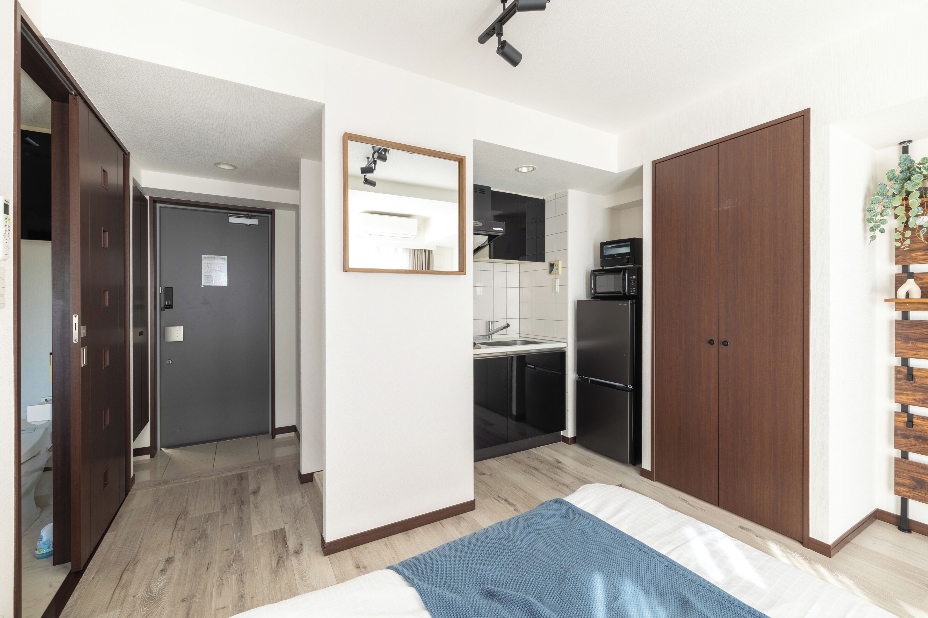 A bedroom with a bed, a wooden wardrobe, a mirror, and a kitchenette in the background.