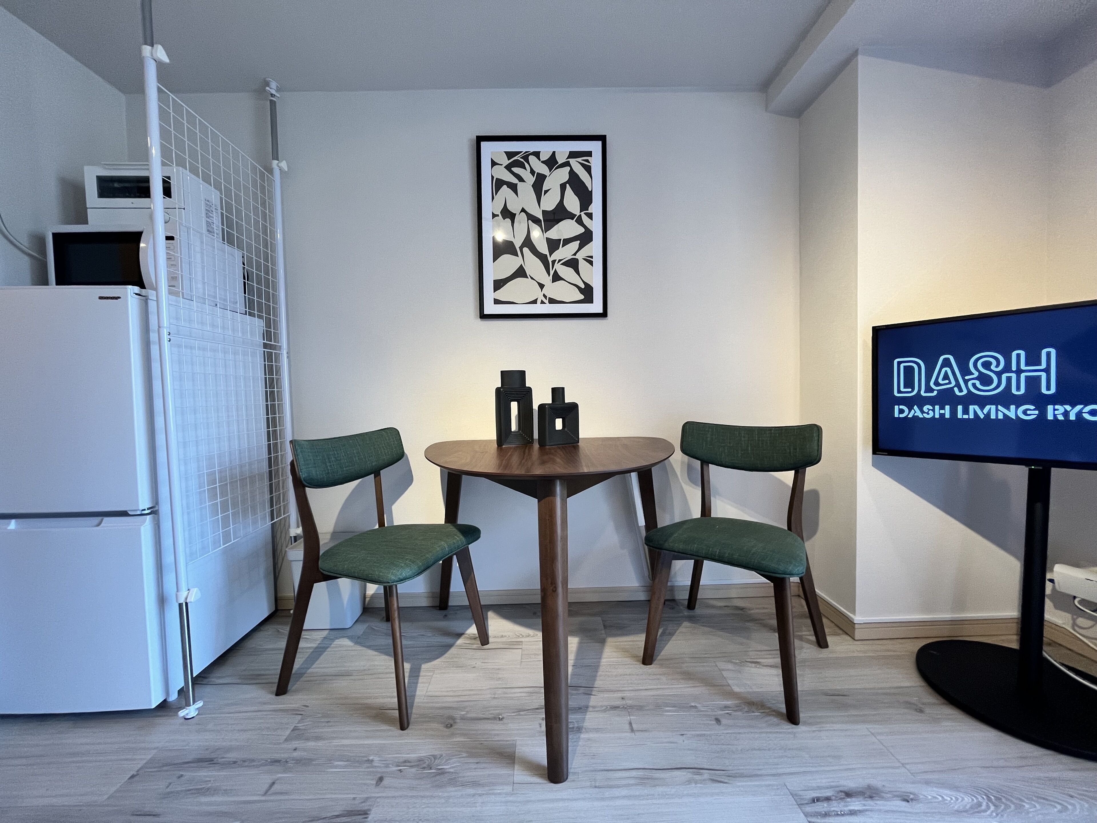 A modern kitchen with a white refrigerator, a microwave, and a coffee machine. There is a dining table with two chairs and a framed artwork on the wall.