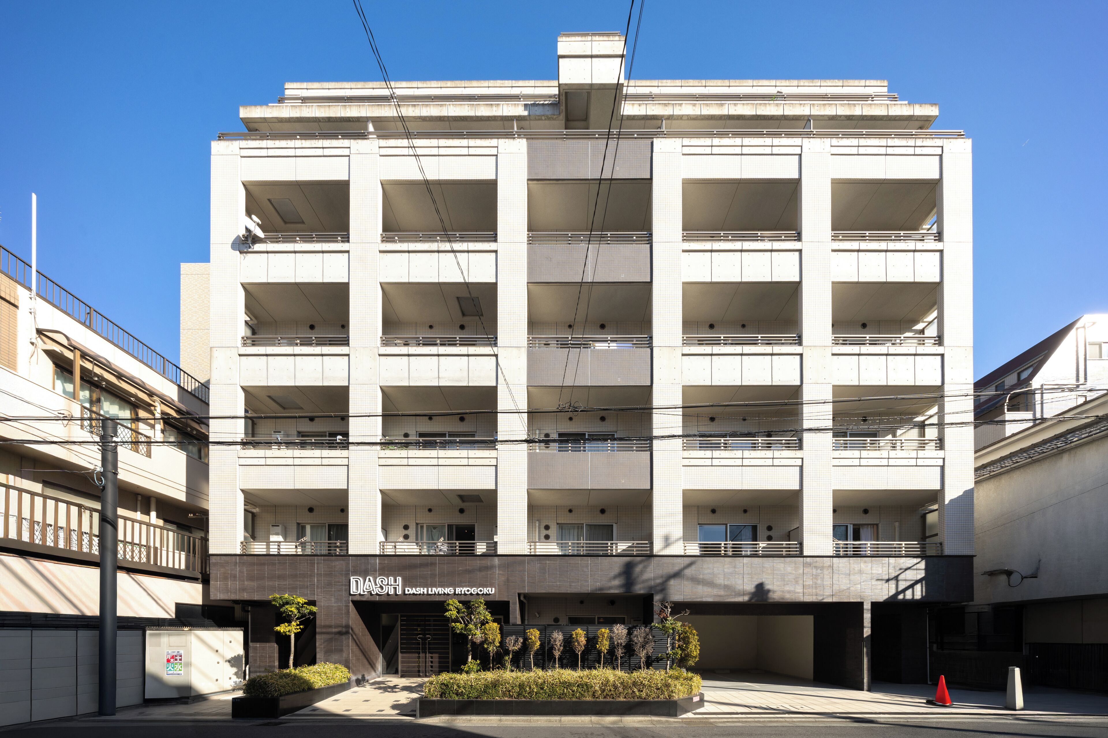 Un edificio de varios pisos con balcones, un letrero que dice 'DASH DASH LIVING ITOGOKU' y un pequeño jardín delantero.