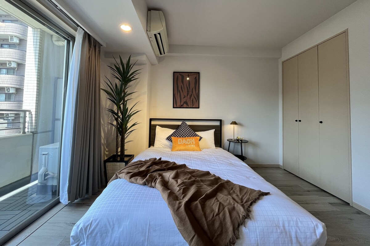 A bedroom with a bed, a potted plant, a nightstand, a lamp, a window with curtains, and a view of buildings outside.