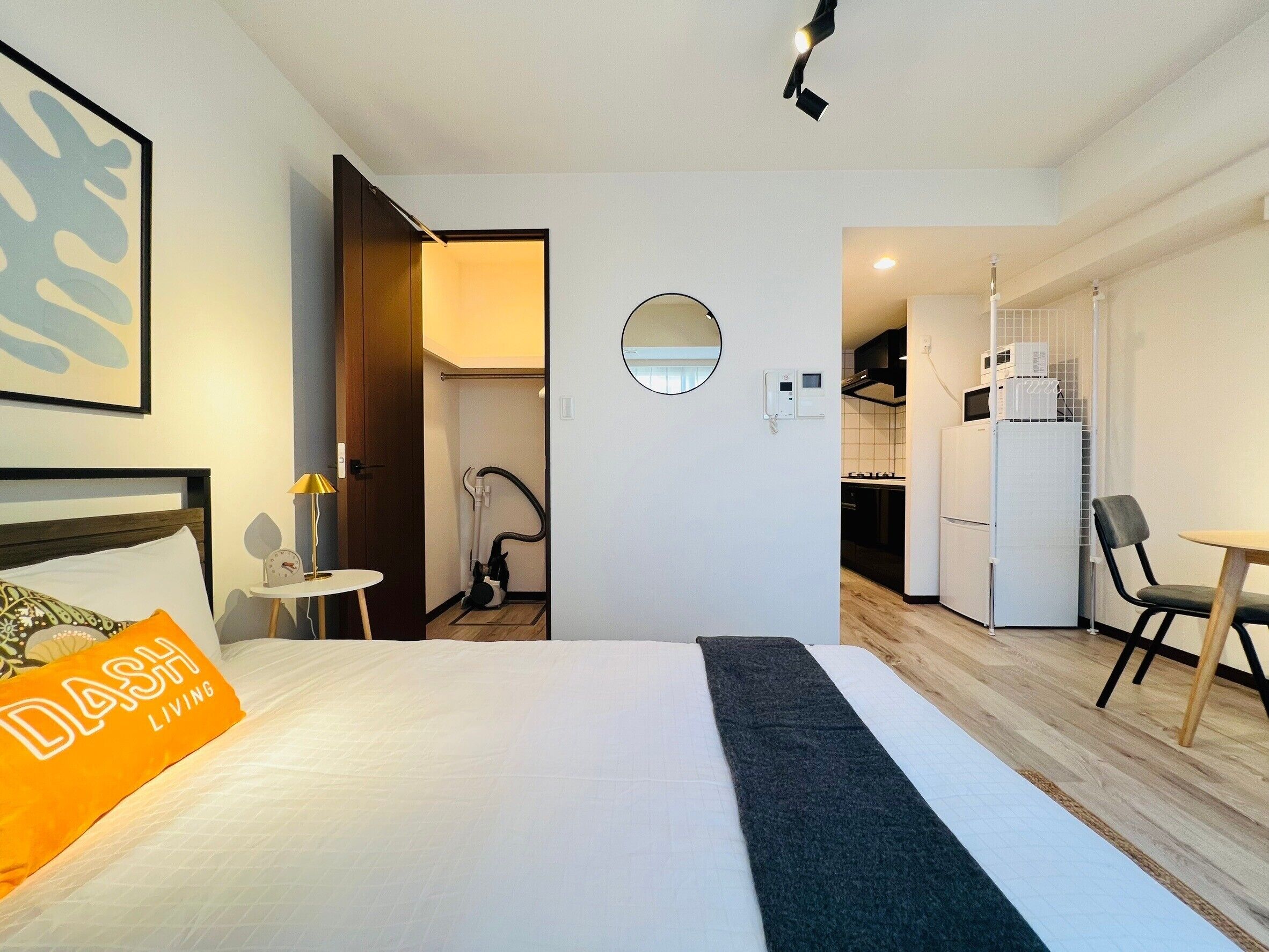 A hotel room with a bed, a desk, a chair, a small table, a mirror, and a kitchenette.