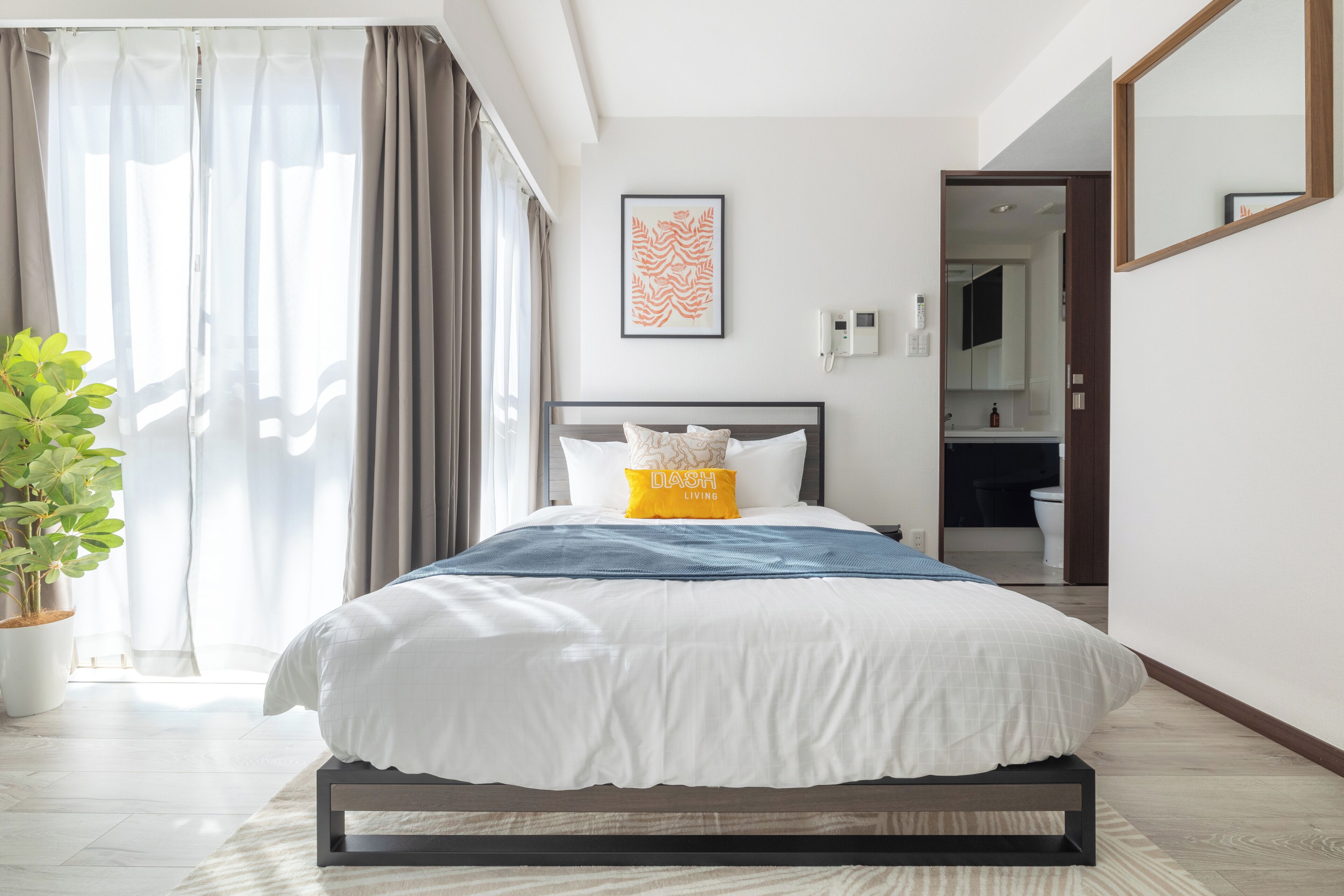 A modern hotel room with a large bed, a bedside table, a potted plant, a bathroom visible through an open door, and a framed artwork on the wall.