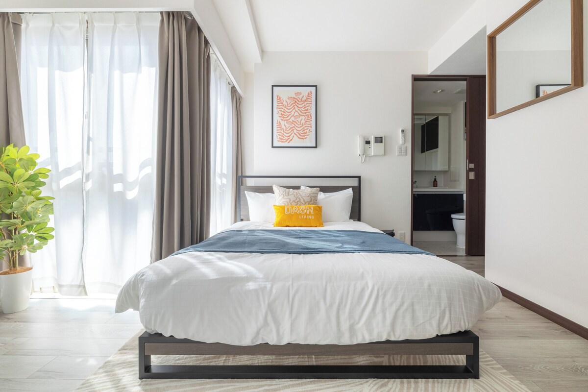A modern hotel room with a large bed, a bedside table, a potted plant, a bathroom visible through an open door, and a framed artwork on the wall.