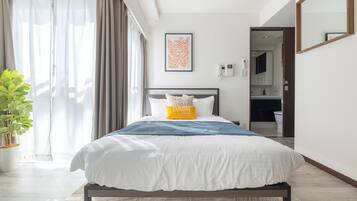 A modern hotel room with a large bed, a bedside table, a potted plant, a bathroom visible through an open door, and a framed artwork on the wall.