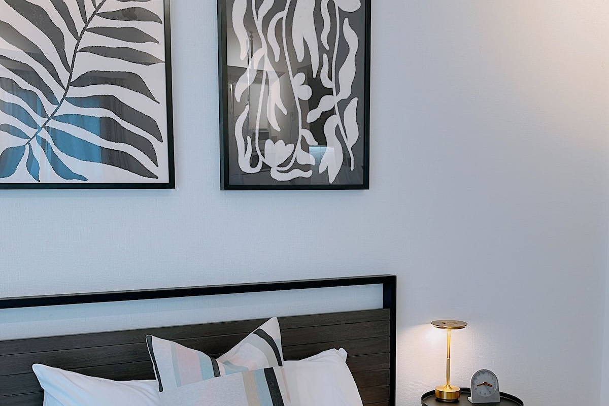 A neatly made bed with a blue throw blanket, a bedside table with a lamp, and two abstract wall art pieces.