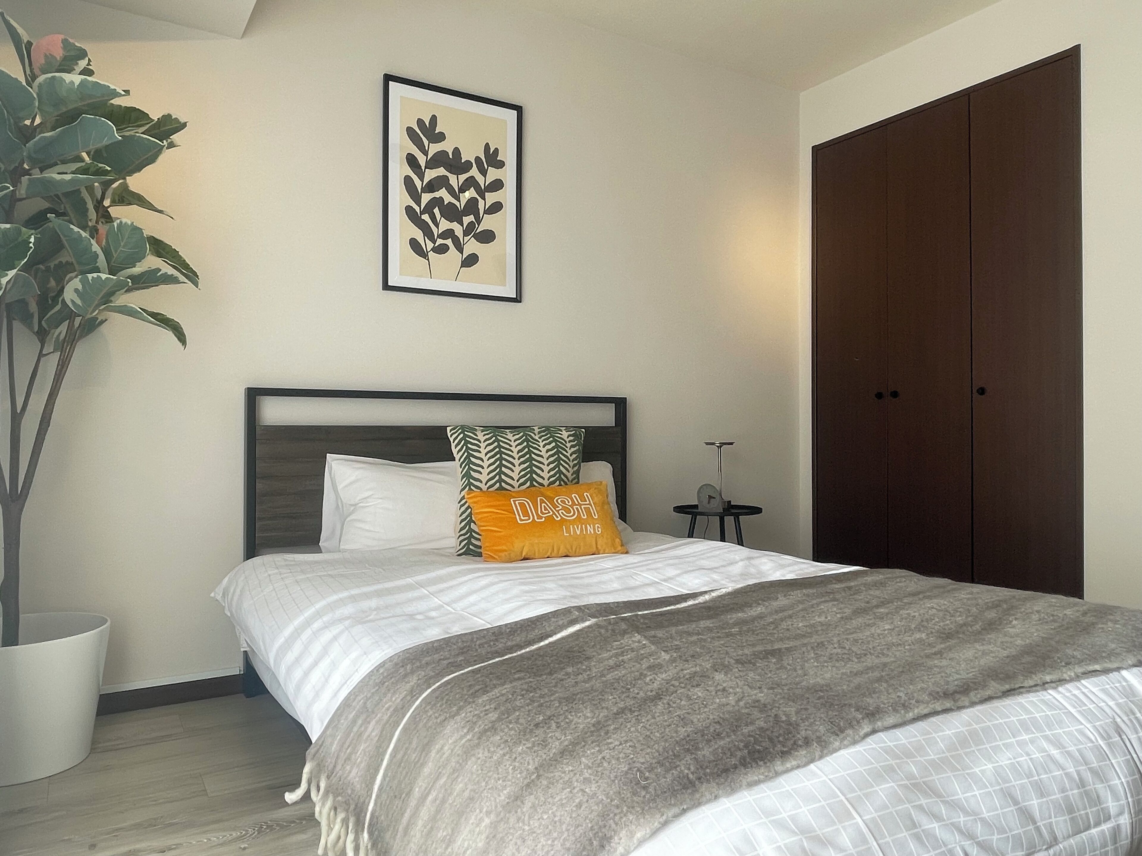 A bedroom with a bed, a bedside table, a potted plant, a wardrobe, and a framed artwork on the wall.