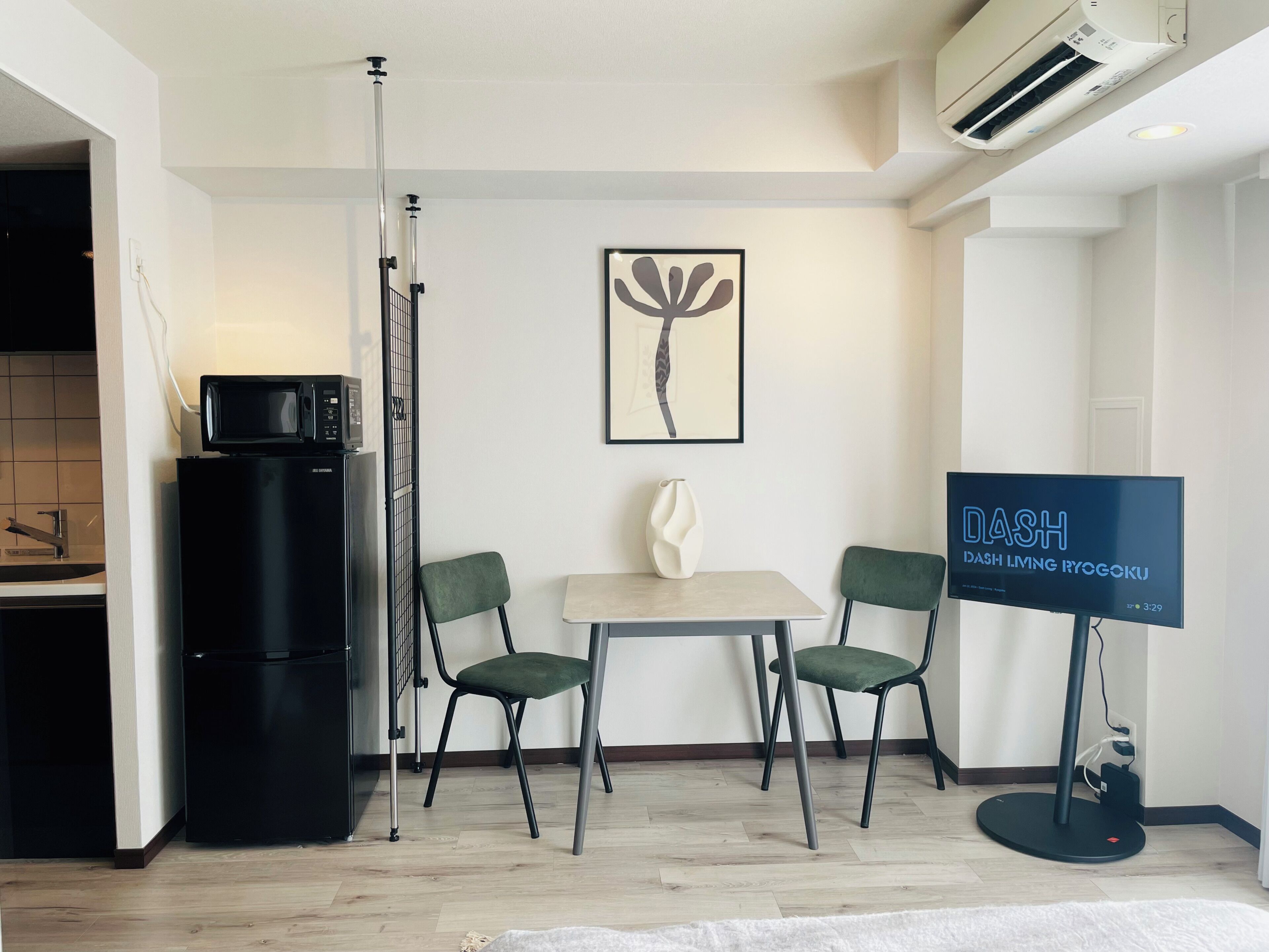 A modern room with a dining table, two chairs, a black refrigerator, a wall-mounted air conditioner, and a flat-screen TV displaying 'DASH'.