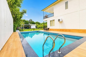 Une piscine avec des échelles en métal, une terrasse en bois et un bâtiment blanc doté d’un balcon.