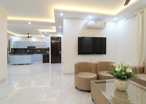 A modern living room with a flat-screen TV, a sofa, a coffee table, and a kitchen in the background. - 4 Bedroom Farmhouse With Private Pool In Noida (Nodia)