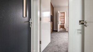 Un passadís amb una porta fosca, una porta blanca i una porta de fusta que condueix a una habitació amb una paret blanca i una prestatgeria.