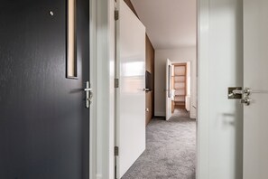 A hallway with a dark door, a white door, and a wooden door leading to a room with a white wall and a shelf. - Marine Place | Flat 5 (St Andrews)