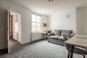 Een kamer met een grijs vloerkleed, een grijze bank, een houten tafel, een raam en een radiator.