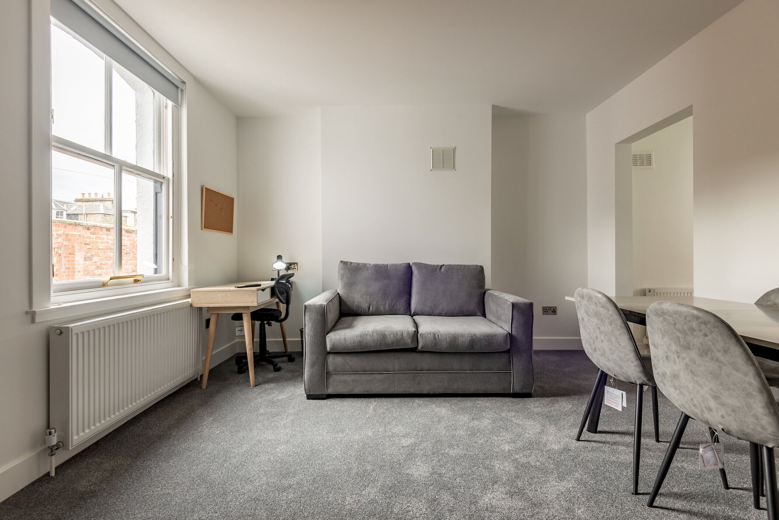 A room with a grey sofa, a small wooden table, a chair, and a window with a view of buildings.