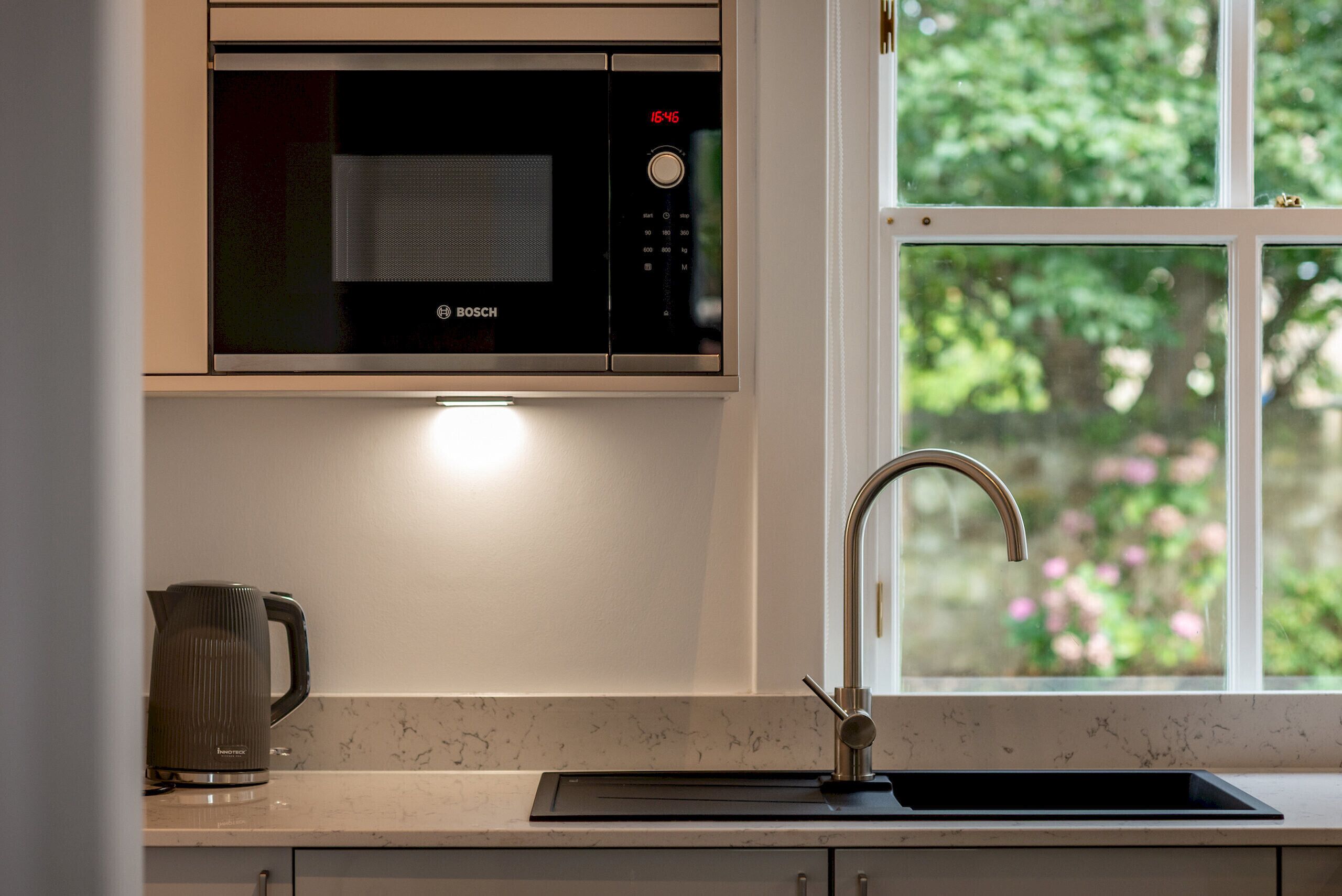 A kitchen with a Bosch microwave, a kettle, a sink, and a window with a view of greenery.