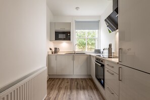 A compact kitchen with white cabinetry, a built-in microwave, a window with a view, and a white radiator. - Marine Place | Flat 5 (St Andrews)