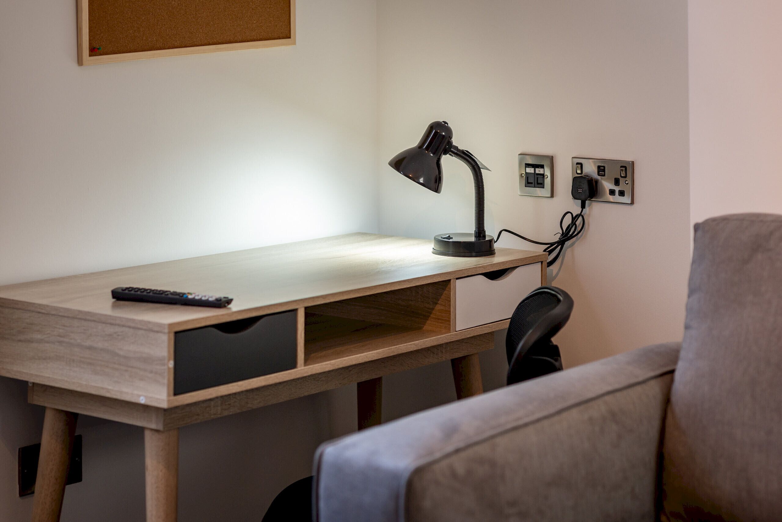 A desk with a lamp, a remote control, and a power outlet.