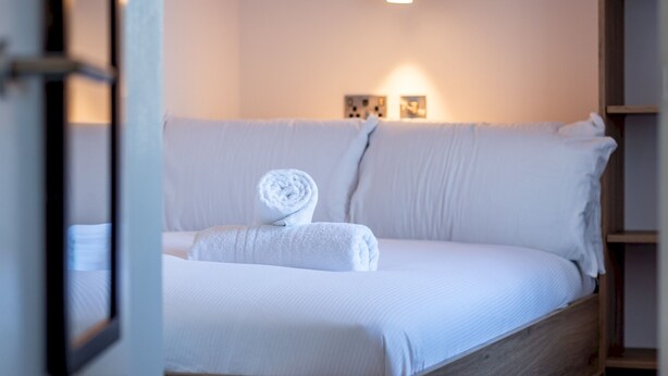 Un lit bien fait, avec du linge de lit blanc et une serviette enroulée, une tête de lit en bois et un luminaire fixé au mur.