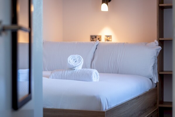 A neatly made bed with white linens and a rolled towel, a wooden headboard, and a wall-mounted light fixture. - Marine Place | Flat 11 (St Andrews)