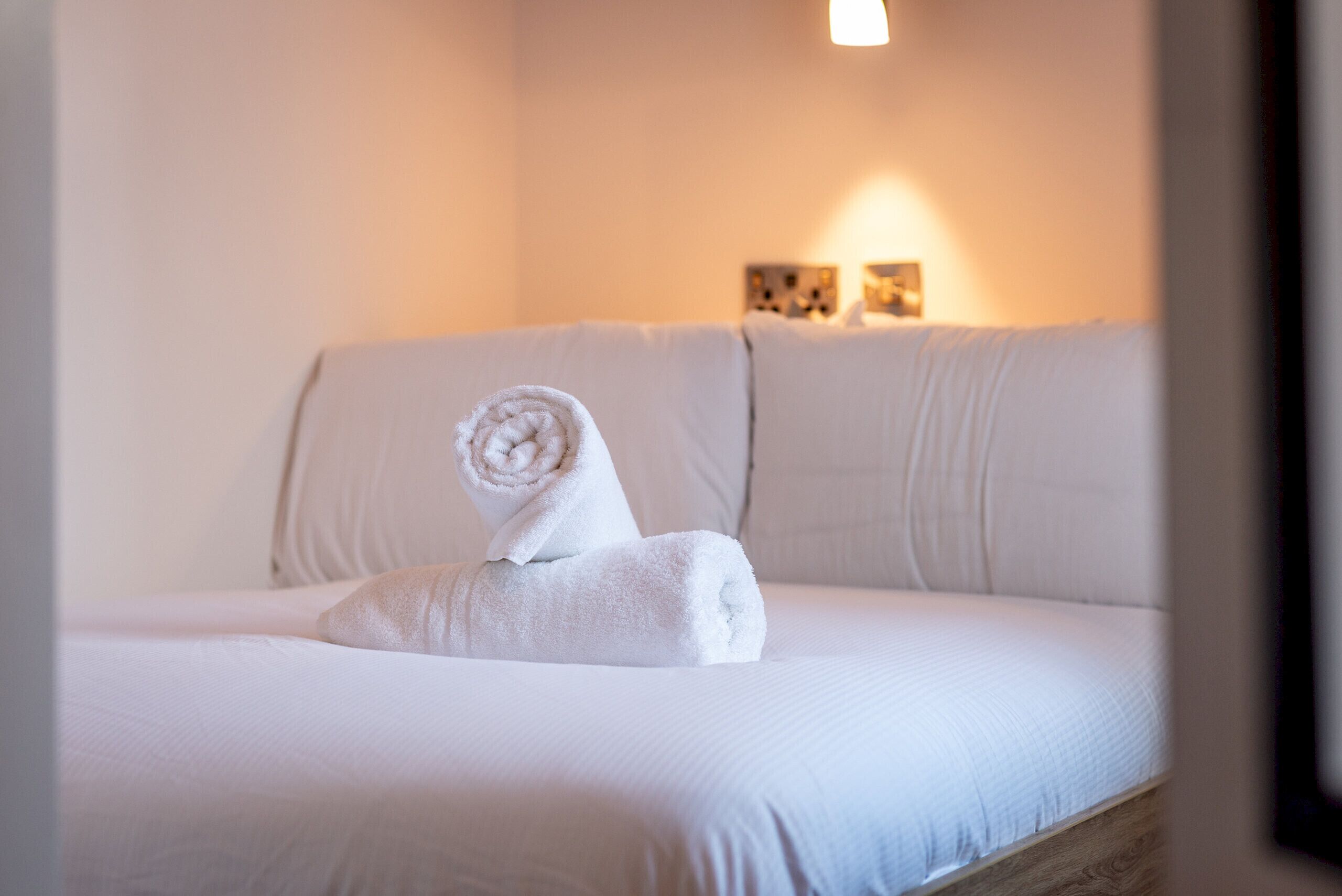 A neatly made bed with a rolled towel, a light-colored sofa, and a wall-mounted light fixture.