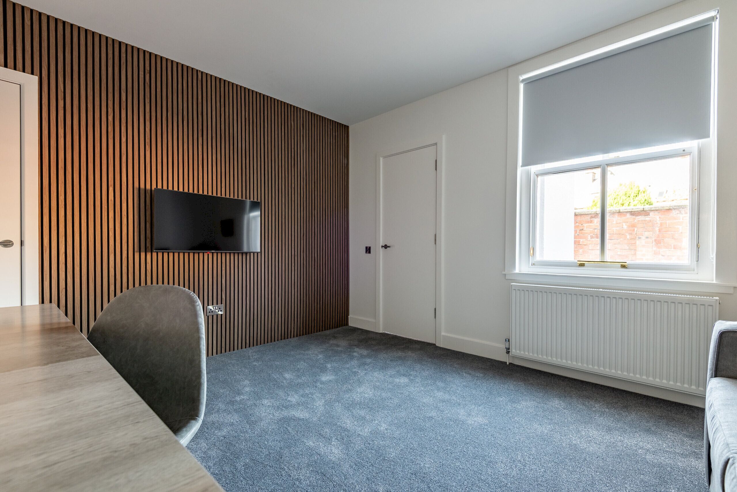 Una habitación moderna con un panel de madera en la pared, un televisor de pantalla plana, un ventanal grande y una alfombra gris.