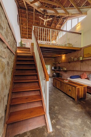 A wooden staircase leading to a loft area with a ceiling fan, a kitchen with a refrigerator, and a dining area with a bench.