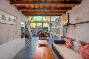 Une piĂšce Ă©troite avec une table en bois, un canapĂ© ornĂ© de coussins Ă motifs, un mur en pierre et une fenĂȘtre donnant sur un paysage verdoyant.