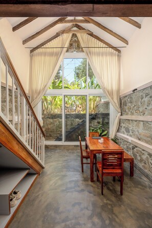 A dining area with a wooden table and chairs, a staircase, and a view of greenery through large windows.