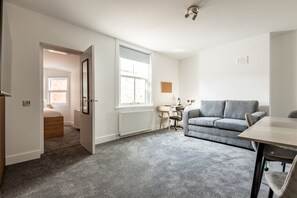 Ein Zimmer mit grauem Sofa, Schreibtisch aus Holz, Stuhl und Heizkörper.