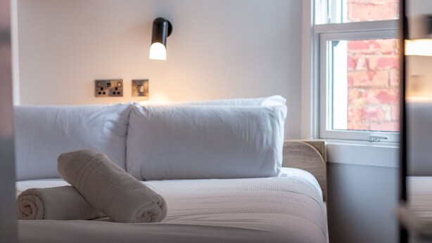 Un lit bien fait, avec une serviette enroulée, un luminaire fixé au mur et une fenêtre donnant sur un mur de briques.