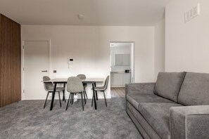 Ein modernes Wohnzimmer mit einer grauen Couch, einem Esstisch mit Stühlen und einem durch eine offene Tür sichtbaren Küchenbereich.
