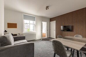 Ein modernes Zimmer mit einer grauen Couch, einem Holzesstisch mit Stühlen, einem an der Wand montierten Fernseher und einem Fenster mit Außenblick.