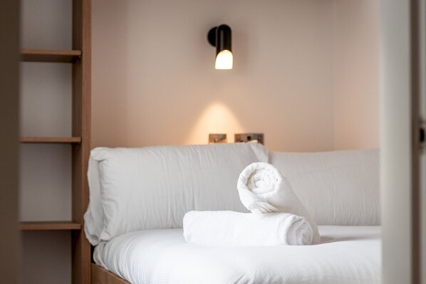 A neatly made bed with rolled towels, a wall-mounted light fixture, and a wooden shelf unit. - Marine Place | Flat 4 (St Andrews)