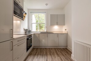 A modern kitchen with white cabinetry, a Bosch microwave, a double oven, and a window providing natural light. - Marine Place | Flat 4 (St Andrews)