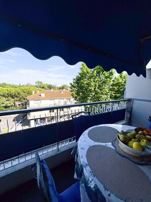 Un balcón con una mesa y sillas, vistas a edificios y árboles, y un toldo azul.