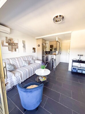 A modern living room with a sofa, a blue armchair, a small round table, and a kitchen in the background.