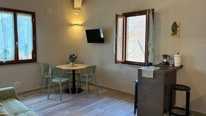Une petite pièce bien éclairée, avec une table ronde, deux chaises, un téléviseur fixé au mur et une fenêtre donnant sur un immeuble en briques.