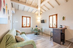 A room with a sofa, a dining table with chairs, a small kitchen area with a bar stool, and a TV mounted on the wall. - Il Cappero Apartment in the Center of Buonconvento (Buonconvento)
